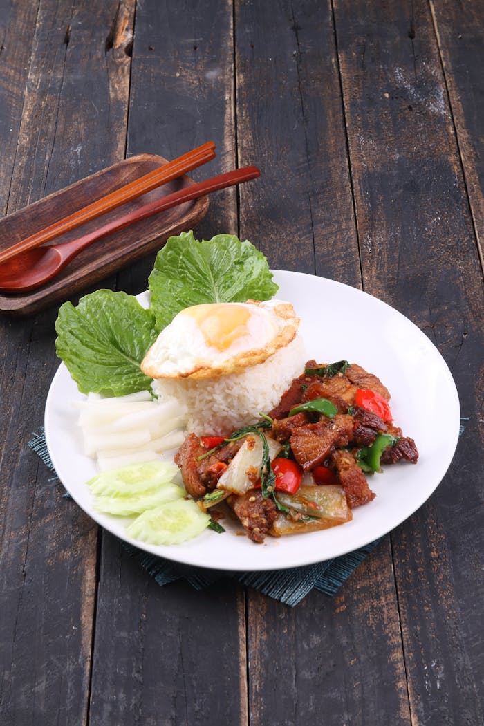 Thai basil chicken with rice, fried egg, and fresh vegetables on a rustic wooden table.