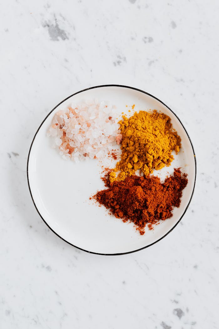 Top view of different powdered spices of curcuma and chili with pink salt in metal bowl on white marble surface